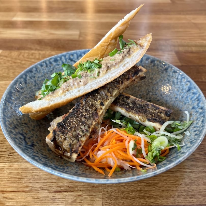 Grilled bone marrow with a baguette, topped with a creamy spread, cilantro, and pickled vegetables.