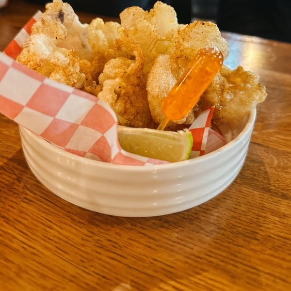 Crispy fried chicharrones in a white bowl with a lime wedge and hot sauce on a wooden table.