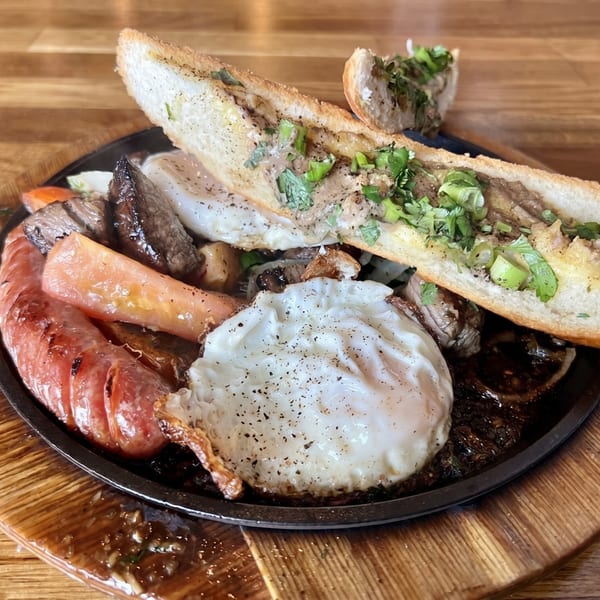 Sizzling platter with sausage, fried eggs, steak, and a baguette topped with herbs.