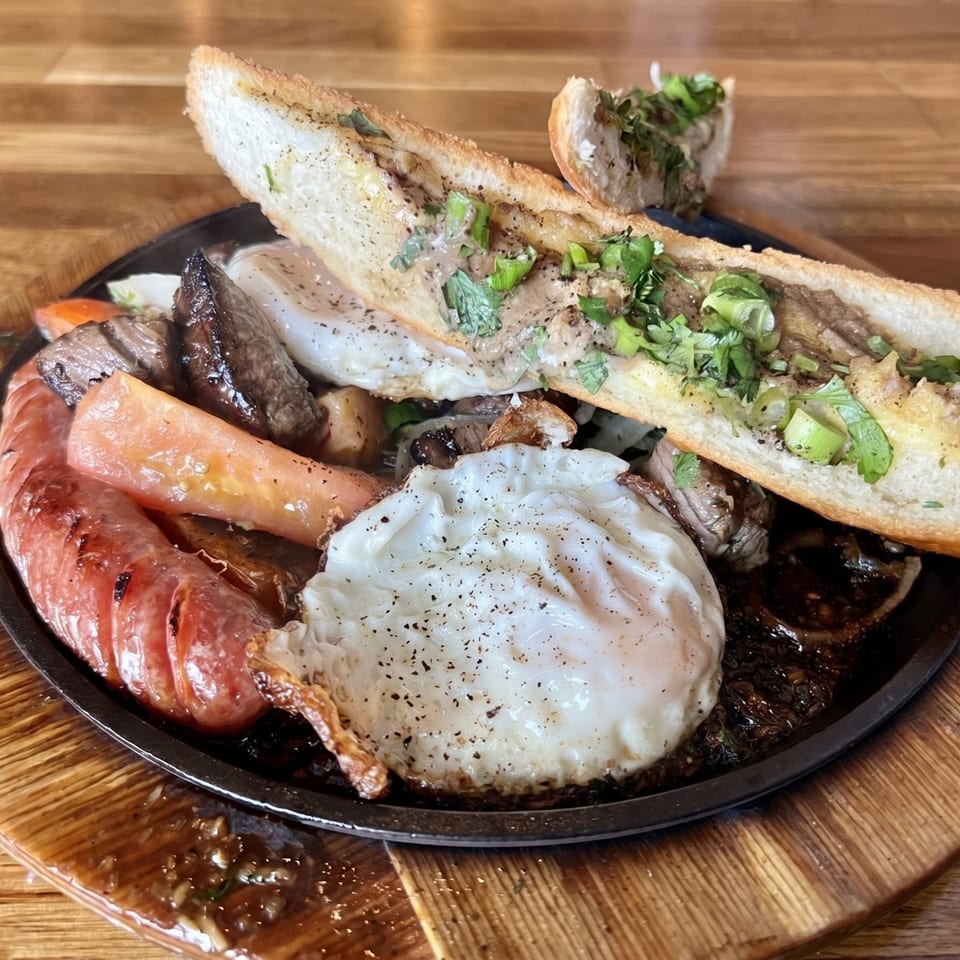 Sizzling platter with sausage, fried eggs, steak, and a baguette topped with herbs.
