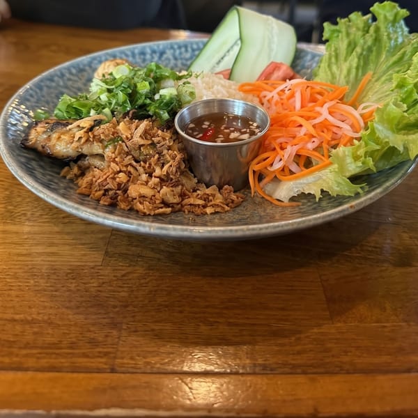 A colorful dish with grilled meat, crispy fried onions, rice, fresh vegetables, and a dipping sauce.